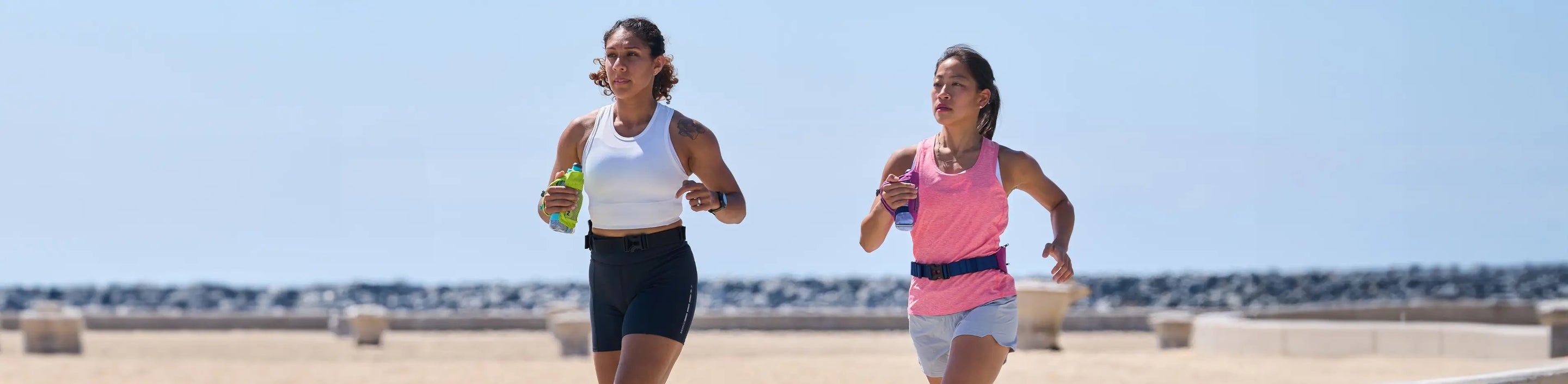 two women running at the beach while wearing Nathan Sports tank tops in white and pink